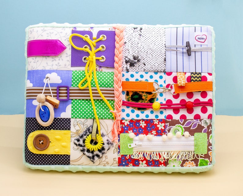 May include: A multi-colored fabric activity mat with various textures and features for babies and toddlers. It includes a zipper, buttons, laces, a tag, and a variety of fabrics.