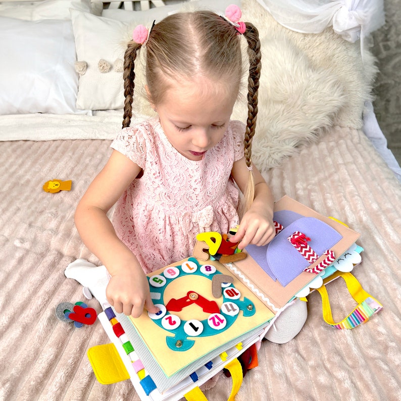以下が含まれることがあります： 子供がカラフルなフェルトの活動ブックで遊んでいます。この本は、動く針のある時計の文字盤、紫色のフェルトのポケット、そしてさまざまなテクスチャとアクティビティを特徴としています。