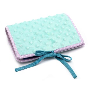 Puede incluir: Un libro de tela azul claro y morado con un lazo turquesa. El libro tiene una cubierta suave y texturizada y es perfecto para un beb&eacute; o un ni&ntilde;o peque&ntilde;o.