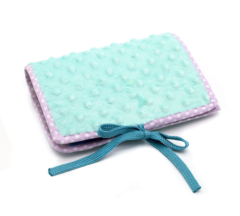Puede incluir: Un libro de tela azul claro y morado con un lazo turquesa. El libro tiene una cubierta suave y texturizada y es perfecto para un beb&eacute; o un ni&ntilde;o peque&ntilde;o.