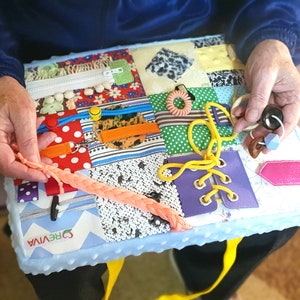May include: A blue and white sensory activity mat with various textures and objects, including a zipper, buttons, laces, and a hair tie. The mat is labeled "CREVIVA" in white letters.