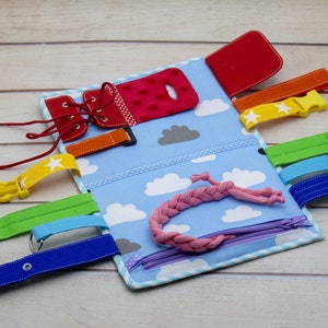 Puede incluir: Un libro de actividades de tela azul y blanco con varias texturas y cierres para ni&ntilde;os. Cuenta con un zapato rojo con cordones, una cremallera, una hebilla, una correa de velcro, un bot&oacute;n y un cord&oacute;n rosa trenzado.