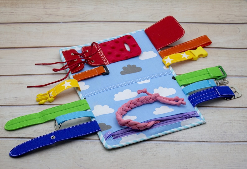 Puede incluir: Un libro de actividades de tela azul y blanco con varias texturas y cierres para ni&ntilde;os. Cuenta con un zapato rojo con cordones, una cremallera, una hebilla, una correa de velcro, un bot&oacute;n y un cord&oacute;n rosa trenzado.