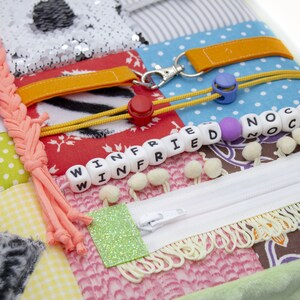 May include: A multi-colored fabric square with various textures and objects attached. The square features a zipper, a braided cord, a yellow rubber band, a key chain, a small red button, and a string of white beads with the names "WINFRIED" and "NOCK" spelled out.