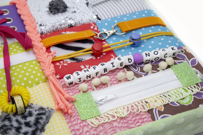 May include: A multi-colored fabric square with various textures and objects attached. The square features a zipper, a braided cord, a yellow rubber band, a key chain, a small red button, and a string of white beads with the names "WINFRIED" and "NOCK" spelled out.