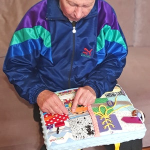 May include: A person is sitting on a couch and interacting with a colorful, multi-textured activity mat. The mat has various textures, including fabric, buttons, zippers, and laces. The mat is blue with a white border and the word "CREWIVA" is printed on it. The mat has a yellow strap.