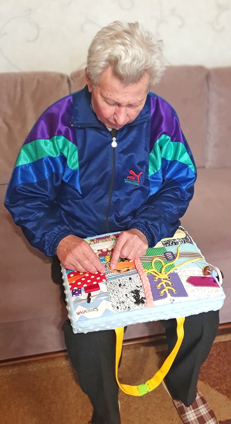 May include: A person is sitting on a couch and interacting with a colorful, multi-textured activity mat. The mat has various textures, including fabric, buttons, zippers, and laces. The mat is blue with a white border and the word "CREWIVA" is printed on it. The mat has a yellow strap.