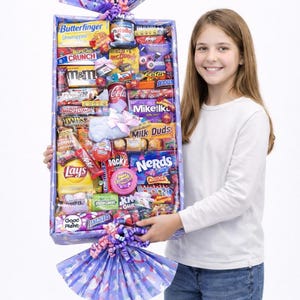 Könnte beinhalten: Eine große rechteckige Geschenkbox gefüllt mit verschiedenen Süßigkeiten, darunter Butterfinger, M&M's und Reese's. Die Box ist mit lila Seidenpapier und Bändern verziert. Eine junge Person hält die Box.