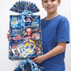 May include: A rectangular gift basket wrapped in clear plastic containing an assortment of snacks including Pepsi, Oreo, Nutella, M&M's, Snickers, and Skittles. The basket is decorated with blue ribbon bows and patterned paper with birthday text.