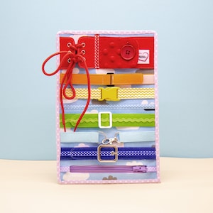 Puede incluir: Un tablero de tela colorido con varias texturas y cierres para que los ni&ntilde;os jueguen. Cuenta con una actividad de encaje roja, una hebilla amarilla, una cremallera verde, un cintur&oacute;n azul con hebilla, un cierre de velcro blanco y una cremallera morada. El tablero tiene un fondo rosa con lunares blancos y nubes blancas.