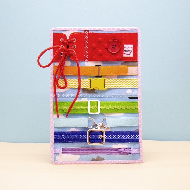 Puede incluir: Un tablero de tela colorido con varias texturas y cierres para que los ni&ntilde;os jueguen. Cuenta con una actividad de encaje roja, una hebilla amarilla, una cremallera verde, un cintur&oacute;n azul con hebilla, un cierre de velcro blanco y una cremallera morada. El tablero tiene un fondo rosa con lunares blancos y nubes blancas.