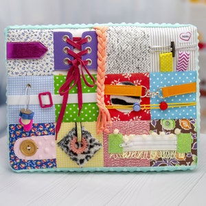 May include: A multi-colored fabric activity book with various textures and features for babies and toddlers. It includes a zipper, buttons, laces, and a variety of fabrics.