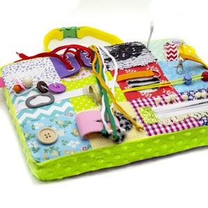 May include: A multi-colored fabric activity mat with various textures and features for babies and toddlers. It includes a zipper, buttons, laces, ribbons, and a tag. The mat is green with white polka dots.