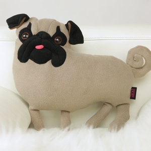 Puede incluir: Un juguete de peluche de perro pug marrón con orejas, nariz y ojos negros. El juguete tiene una cola rizada y está sentado sobre una superficie blanca y esponjosa.