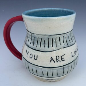 May include: A handmade ceramic mug with a red handle and a light blue interior. The exterior is white with gray bands and vertical lines. The words "You ARE Love" are written on the mug.
