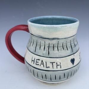 May include: Handmade ceramic mug with a red handle and a light blue interior. The mug features a gray and white design with the word "HEALTH" and a heart symbol. The mug is a unique piece of functional art.