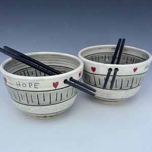 May include: Two ceramic bowls with black chopsticks. The bowls are off-white with a gray stripe design and red heart accents. One bowl has the word "HOPE" written on it. The other bowl has the word "HAPPY" written on it.