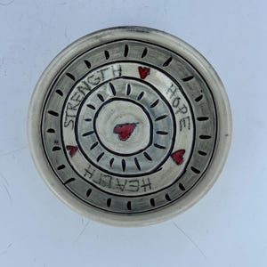 May include: A handmade ceramic dish with a circular design. The dish is primarily off-white with a gray border and accents. The words "STRENGTH", "HOPE", and "HEALTH" are written around the center, with red heart accents.