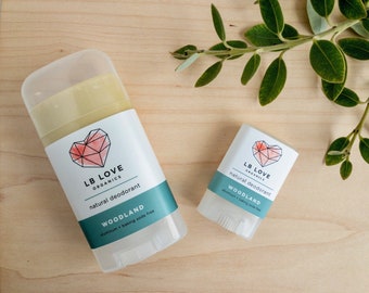 May include: Two deodorant sticks, one larger and one smaller, both with a white label and a pink heart logo. The label reads "LB LOVE ORGANICS natural deodorant WOODLAND aluminum + baking soda free".
