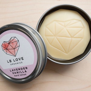 Pode incluir: Uma lata prateada com um rótulo rosa e branco que diz "LB LOVE ORGANICS LAVENDER VANILLA hard lotion". O rótulo apresenta um design de coração geométrico rosa. A lata contém uma barra de loção dura branca em forma de coração com um design geométrico.