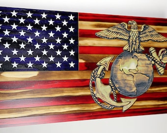 May include: A wooden American flag with a distressed finish. The flag features a blue field with white stars and red and white stripes. The Marine Corps emblem is displayed in the lower right corner of the flag. The emblem is made of metal and features a globe, an anchor, and an eagle.