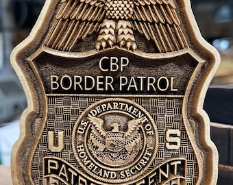 May include: A wooden cutout of a U.S. Customs and Border Protection (CBP) badge. The badge features an eagle with outstretched wings, the text "CBP BORDER PATROL", and the seal of the U.S. Department of Homeland Security.