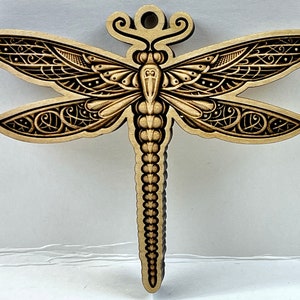May include: A wooden dragonfly ornament with intricate laser-cut details. The dragonfly is brown with black accents and has a long, slender body.