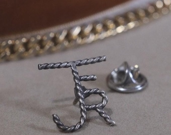 May include: A silver-toned lapel pin featuring the intertwined letters "J" and "R" in a twisted rope design. The pin has a matching backing for secure attachment. The background is a neutral beige.