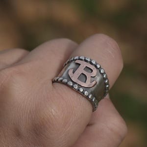 May include: A silver ring with a textured band and a copper letter "B" in the center.