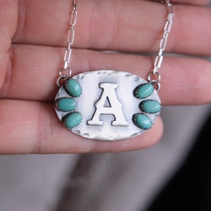 May include: Silver-toned necklace with an oval pendant featuring the letter "A". The pendant is adorned with six turquoise-colored stones and hangs from a delicate chain. The pendant has a hammered texture.
