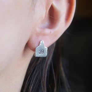 May include: Silver earrings shaped like a livestock ear tag with the number 39 engraved on the front.