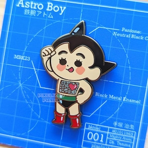 May include: A black metal enamel pin featuring Astro Boy, a Japanese manga character. The pin is on a blue background with a grid pattern. The text "Astro Boy" and "鉄腕アトム" are printed on the background. The pin is a collectible item for fans of anime and manga.