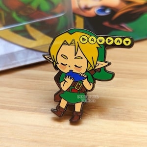 Puede incluir: Un pin de esmalte de color dorado que representa un dibujo animado de Link de la serie de videojuegos The Legend of Zelda. Link lleva su característica túnica y gorro verdes y está tocando la Ocarina del Tiempo. El pin tiene un esquema de color azul y verde y presenta el texto "Pixellized Panda" en la parte inferior.