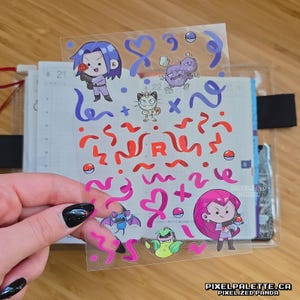 May include: Transparent sticker sheet with cartoon characters and decorative elements. The sheet includes images of characters, hearts, ribbons, and the letter "R" in red and pink. The sticker sheet is held over a planner.