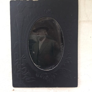 May include: Antique black framed portrait of a man in a suit and hat. The oval portrait is surrounded by an embossed floral design. The frame has an embossed design on the left side.