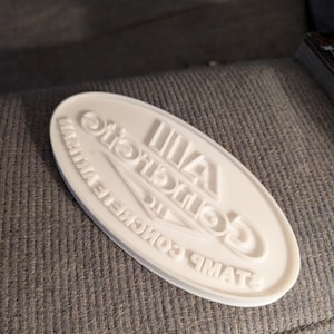 May include: A white, oval-shaped stamp with the text "All Concrete Stamp Inc" in a circular arrangement. The stamp is made of a solid material and is designed for imprinting concrete.