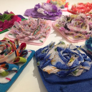 May include: Assortment of colorful fabric flower brooches, each with a unique design and texture. The brooches are made with different fabrics, including lace, tulle, and felt. They are perfect for adding a touch of whimsy to any outfit.