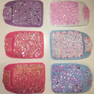 May include: Six small, rectangular, glittery felt patches in various colours, including purple, pink, light blue, and pink with a white border. The patches are all the same size and shape, and they are arranged in two rows of three.