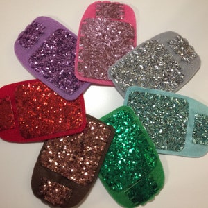 Op de afbeelding: Zeven viltpatches met verschillende glitterkleuren, elk met een gestikt rechthoek in het midden. De patches zijn in een cirkel gerangschikt.