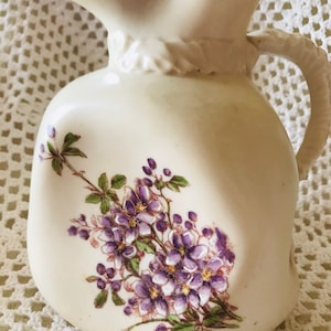May include: A white ceramic pitcher with a floral design. The pitcher has a wide mouth and a handle. The floral design features purple flowers and green leaves.