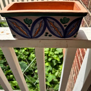 May include: A rectangular terracotta planter with a white glazed exterior featuring hand-painted blue and orange leaf-like motifs and small green accents. The interior rim is unglazed terracotta, and the top edge is finished with a dark green border.