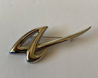May include: A silver-toned brooch with a flowing, abstract design. The brooch features a curved shape with a pointed end, and a pin closure on the back. The jewelry piece is set against a white background.