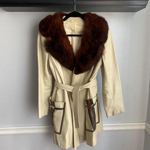 May include: Cream-colored leather coat with a brown fur collar and a matching belt. The coat features two front pockets with green and red striped accents and gold-colored hardware. The coat is hanging on a black hanger.