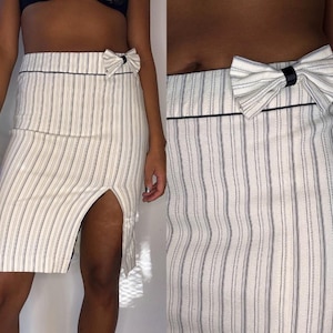 May include: A white pencil skirt with vertical black pinstripes and a front slit. The skirt features a decorative bow at the waist and a black trim along the top. This skirt is a stylish addition to any wardrobe.