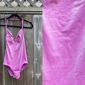 May include: A pink swimsuit with a halter neck and a V-shaped neckline. The swimsuit is hanging on a wooden hanger against a wooden fence.