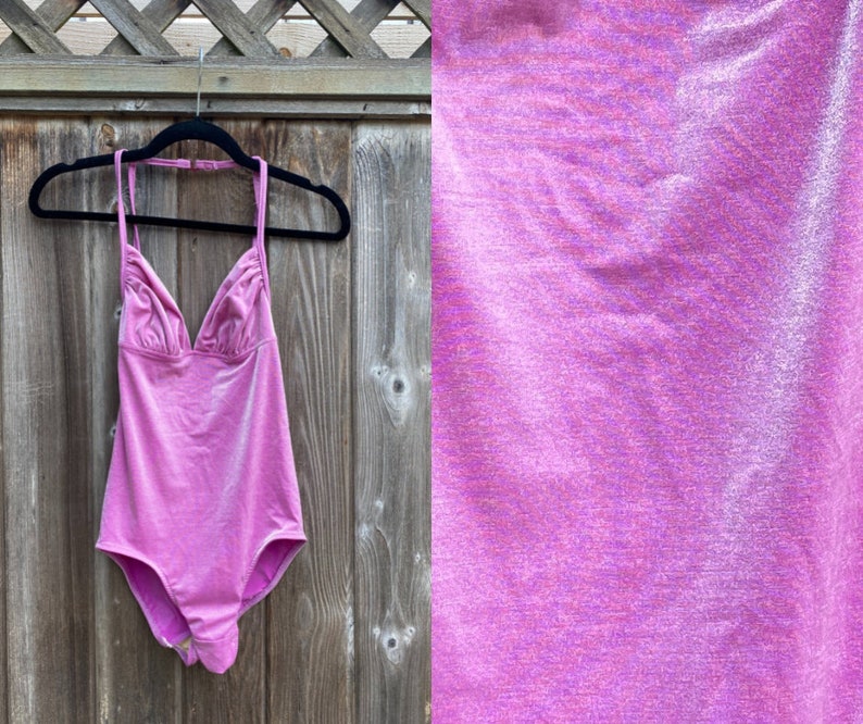 May include: A pink swimsuit with a halter neck and a V-shaped neckline. The swimsuit is hanging on a wooden hanger against a wooden fence.