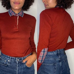 May include: Rust-colored long-sleeve shirt with a plaid collar and elbow patches. The shirt has a button-up placket and is paired with blue denim jeans. The plaid pattern includes shades of blue, red, and green.