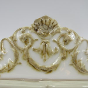 May include: White ceramic decorative piece with a gold-toned, ornate design. The design features intricate swirls and scrolls.