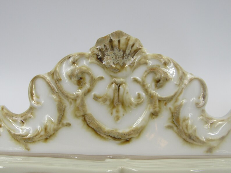 May include: White ceramic decorative piece with a gold-toned, ornate design. The design features intricate swirls and scrolls.