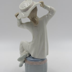 May include: A porcelain figurine of a girl wearing a white dress and a white hat with a pink ribbon. She is sitting on a blue and pink stool.